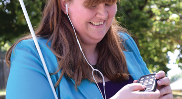 A woman smiles while listening to NFB-NEWSLINE on a Victor Reader Stream and headphones.